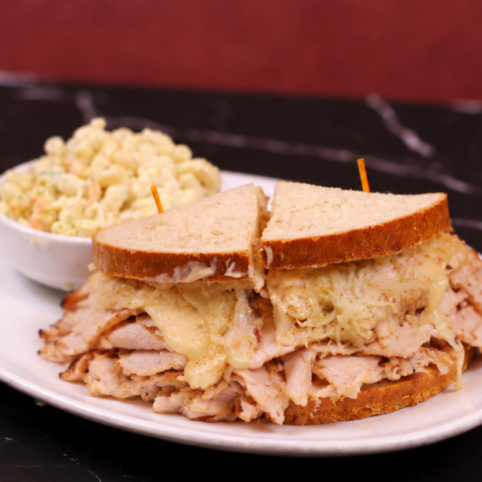 Stacked turkey sandwich with melted cheese on toasted bread, served with a bowl of creamy macaroni salad.