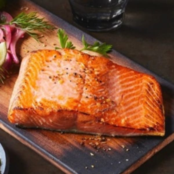 Grilled salmon fillet seasoned with pepper, garnished with dill, red onion, and lime on a wooden board.
