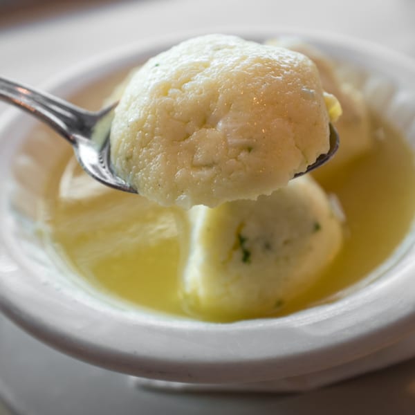 A spoonful of matzo ball soup, with a large matzo ball held above a bowl of broth.