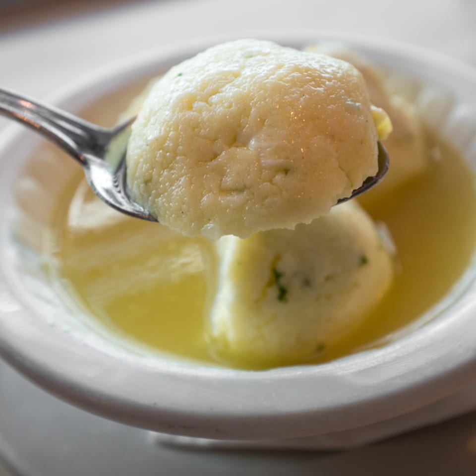 A spoonful of matzo ball soup, with a large matzo ball held above a bowl of broth.