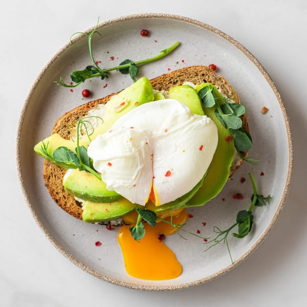Avocado toast with a poached egg, pea shoots, and red pepper flakes on a gray plate.